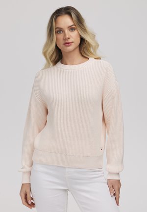 Blondynka w jasnoróżowym dzianinowym swetrze i białych spodniach, stojąca na tle jednolitego szarego tła.