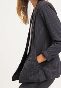 Blazer bleu marine à rayures, avec une coupe ajustée, une fermeture à un bouton et des revers crantés. Le matériau semble lisse avec un léger éclat.