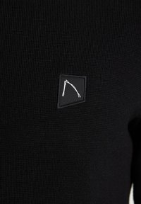 Schwarzer Strickpullover mit einem kleinen schwarzen Patch, der ein weißes abstraktes Design zeigt. Die Textur wirkt gerippt und weich.