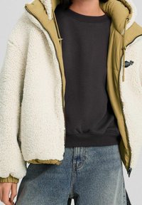 Person iført en beige sherpa fleecejakke med tan-foring over en sort sweatshirt og blå jeans, stående mod en ensfarvet baggrund.