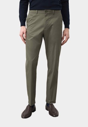 Mężczyzna w oliwkowo-zielonych, dopasowanych spodniach, ciemnych skarpetkach i brązowych skórzanych wsuwanych butach, z rękami luźno opuszczonymi po bokach.