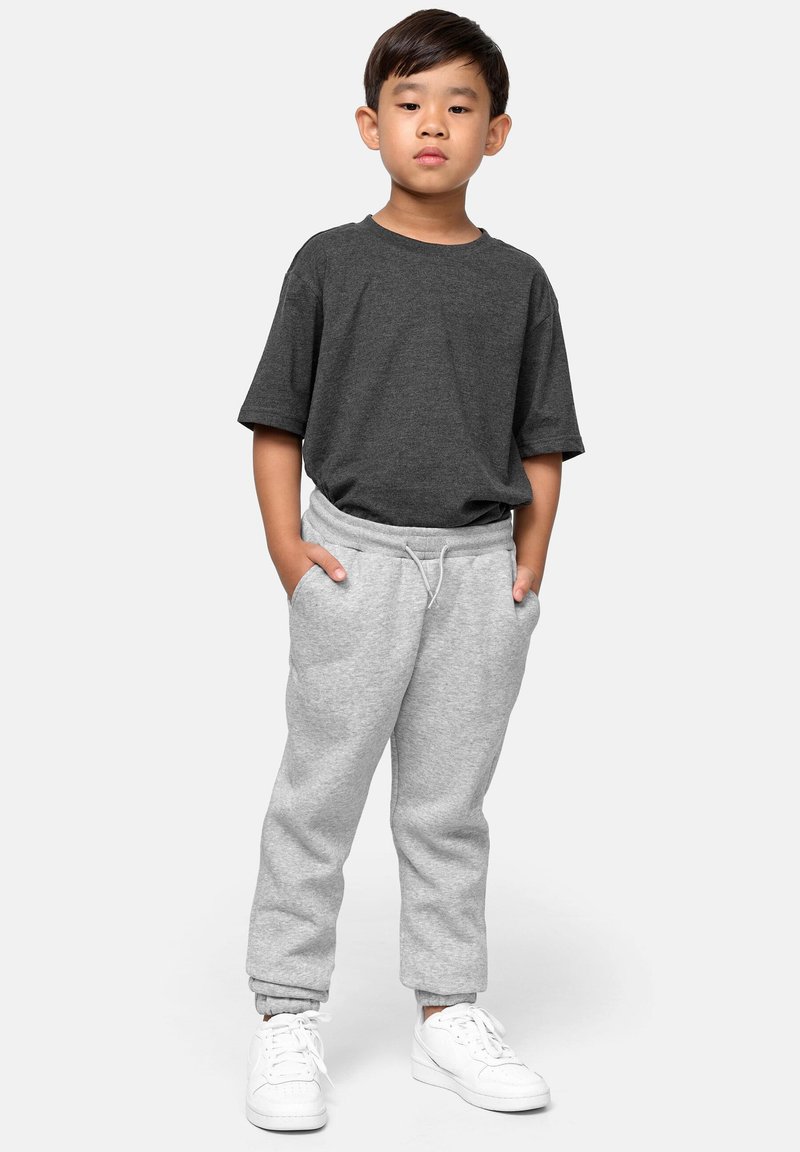 Niño pequeño de pie con las manos en los bolsillos, vistiendo una camiseta gris oscuro, pantalones de chándal gris claro y zapatillas blancas sobre un fondo plano.