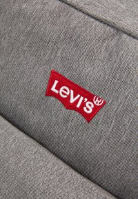Etykieta Levi's na teksturowanej szarej tkaninie, z wyhaftowanym na czerwono logo. Materiał ma subtelny wzór splotu.