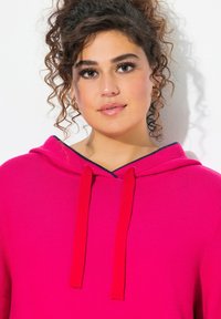 Jasnoróżowa bluza z kapturem o teksturowanym splotem, z czerwoną sznurówką. Posiada kaptur oraz kontrastujący granatowy akcent wzdłuż dekoltu.