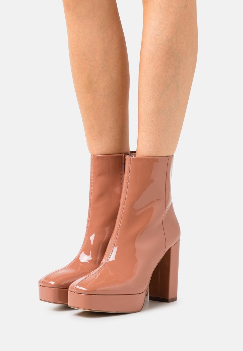 Botines de charol nude brillantes con plataforma y altos tacones de bloque, usados sobre piernas desnudas, contra un fondo blanco.