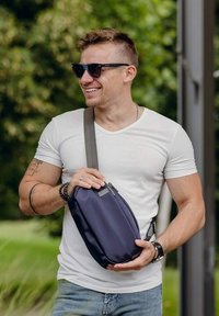Námořnická crossbody taška vyrobená z hladké tkaniny, s zipem a nastavitelným popruhem. Nese ji osoba v bílém tričku a slunečních brýlích.
