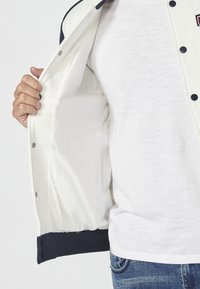 Veste pour homme avec un extérieur crème, des finitions contrastantes en marine et une doublure intérieure blanche. Elle possède des boutons-pression et des poignets côtelés.