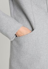 Dłoń częściowo wsunięta w boczną kieszeń jasnoszarego wełnianego płaszcza o gładkiej, teksturowanej tkaninie.