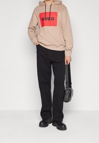 Sudadera beige con un cuadrado rojo y el logo negro "HUGO", combinada con pantalones negros y botas gruesas negras. Los accesorios incluyen un pequeño bolso negro.