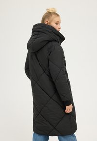 Kobieta o blond włosach związanych w koczek, ubrana w długi, czarny pikowany płaszcz z dużym kapturem, stojąca bokiem na jednolitym tle.