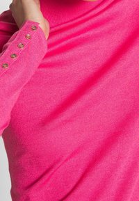 Vartalonmyötäinen vaaleanpunainen neule, jossa on resorikuviointi, poolokaulus ja kultaiset napit hihassa. Pehmeä, joustava kangas.