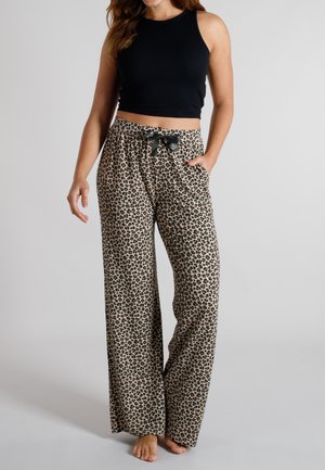 Frau trägt weit geschnittene Leopardenmusterhose mit schwarzer Schleife an der Taille und schwarzes ärmelloses Crop-Top, steht barfuß vor grauem Hintergrund.