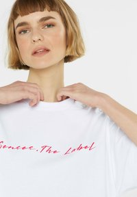 White cotton t-shirt with a crew neck, featuring pink graphic text "Senses.The Label" across the front; smooth texture and relaxed fit.