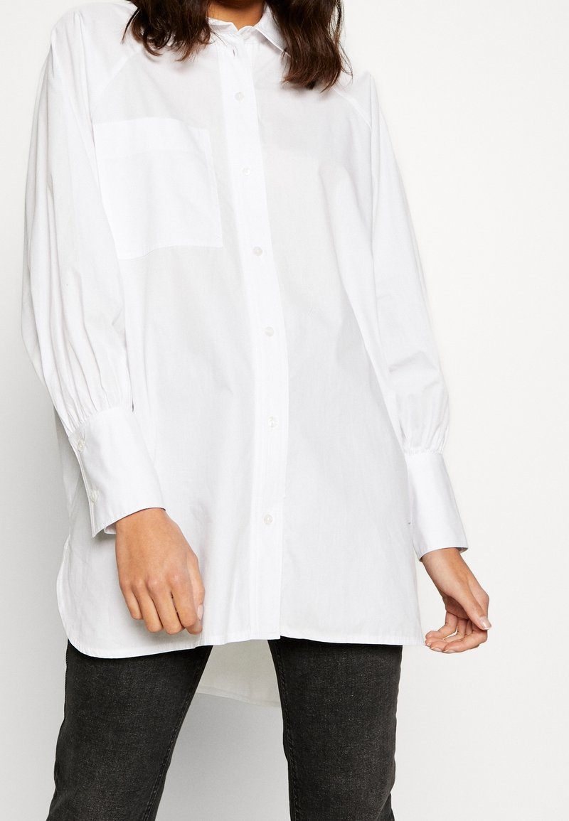 Camicia bianca a maniche lunghe con bottoni realizzata in cotone, dotata di una tasca sul lato sinistro del petto, maniche plissettate e orlo dritto.