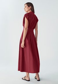 Bordová maxi šaty s krátkymi nafúknutými rukávmi, vysokým golierom a plisovanou sukňou, vyrobené z hladkej matnej látky s priliehavým pásom.