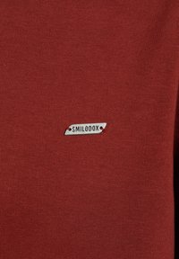 Tessuto rosso con una superficie liscia, dotato di un piccolo etichetta in metallo argentato attaccata con un filo rosso, che reca la scritta "SMILODOX."
