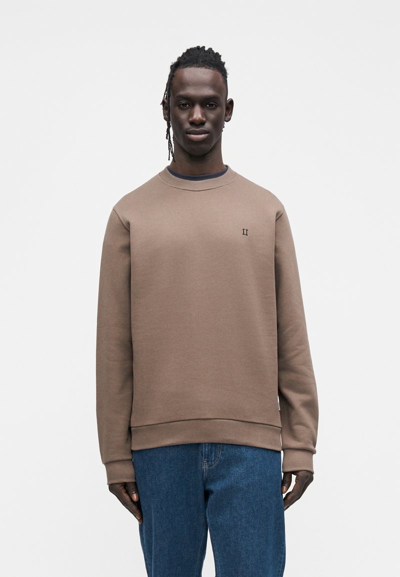 Καφέ φούτερ με στρογγυλή λαιμόκοψη από μαλακό ύφασμα, με ριμπ μανσέτες και τελείωμα, με μικρό λογότυπο στο στήθος. Συνδυάζεται με μπλε τζιν.