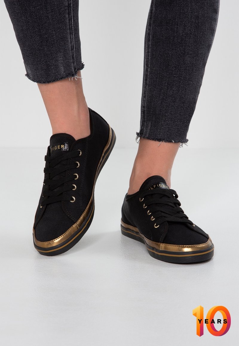 Schwarze Leinenschuhe mit goldenen metallischen Akzenten, flachen Schnürsenkeln und einer glatten Gummisohle. Mit distressed Denim am Knöchel.