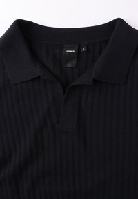 Μαύρο ριμπ μπλουζάκι πόλο με γιακά και κοντή πλακέτα. Διαθέτει υφή υφάσματος και ετικέτα μεγέθους που αναγράφει «L» για μεγάλο μέγεθος.