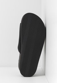 Sandale noire à glissière avec semelle texturée montrant un motif chevron, appuyée contre un mur d’angle blanc sur une surface blanche plane.