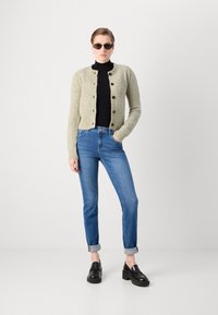 Gebreid vest in lichtgroen met een gestructureerd ruitpatroon, zwarte knopen, gecombineerd met een zwarte coltrui en blauwe jeans, afgemaakt met zwarte instappers.