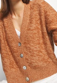 Detail osoby nosící texturovaný tehlově oranžový pletený kardigan s velkými hnědými knoflíky přes bílé tričko.