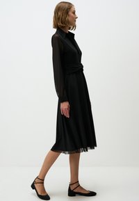 Abito nero con maniche lunghe trasparenti, dettaglio in vita avvolgente, gonna svasata e lunghezza sotto il ginocchio. Indossato con scarpe flat nere.