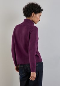 Maglione a collo alto viola con schiena testurizzata, orlo a costine e maniche lunghe. Indossato con jeans in denim scuro, che mettono in evidenza uno stile aderente.