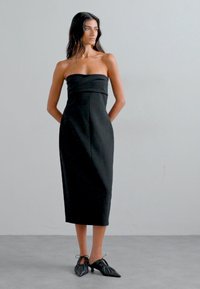 Schwarzes, trägerloses Midikleid mit taillierter Silhouette und geradem Schnitt. Das Model trägt schwarze Stiefeletten mit spitzen Zehen und Schnürung.