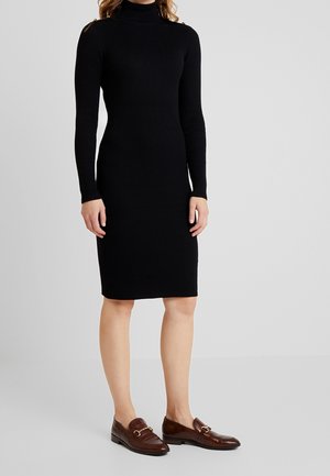 Kobieta ubrana w czarną prążkowaną sukienkę z golfem i długimi rękawami oraz brązowe skórzane mokasyny ze złotymi klamrami, stojąca na tle jednolitego tła.