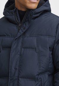 Jachetă puffer albastru închis, cu glugă, fermoar frontal, nasturi presati și textura matlasată cu detalii vizibile de cusătură.