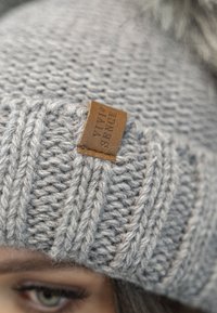 Gray knitted beanie with a textured pattern, featuring a brown leather label on the front. The material appears soft and thick.