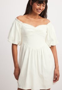 Biela šaty bez ramienok s nafúknutými rukávmi, nariadenou trupovou časťou a rozšírenou sukňou. Mäkko tkanina s jemnou textúrou a uvoľneným strihom.