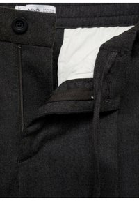 Pantalon gris foncé avec braguette partiellement ouverte à fermeture éclair et bouton noir, montrant la doublure blanche de la poche intérieure et l’étiquette de la ceinture.