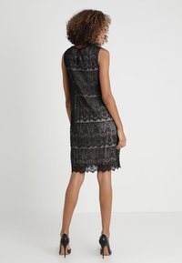 Robe en dentelle noire avec une silhouette cintrée, un design sans manches, un ourlet festonné et un détail dos en forme de goutte. Associée à des talons hauts noirs.