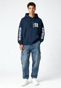 Marineblauwe hoodie met witte opdruk op de borst en letters op de mouwen, gecombineerd met lichtblauwe cargobroek en witte sneakers.