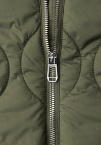 Close-up of a silver zipper on olive green quilted fabric with curved stitching detail.