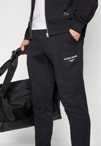 Čierne tepláky vyrobené z mäkkej bavlny, so bočnými vreckami a bielym logom. Dizajn zahŕňa úzky strih a hladkú textúru.