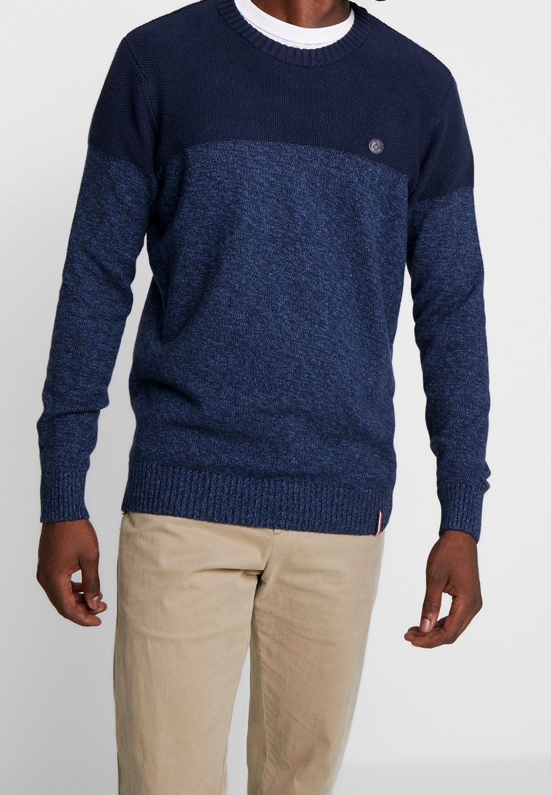 Homme portant un pull à col rond bicolore bleu marine et pantalon beige, debout devant un fond clair uni.