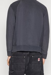 Sweat-shirt gris foncé avec ourlet et poignets côtelés, présentant une texture lisse et des manches raglan. Associé à un pantalon en denim noir.