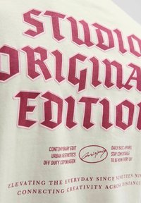 Balts kokvilnas T-kreklis ar lieliem tumši sarkaniem un rozā burtiem. Teksts ietver "STUDIO ORIGINAL EDITION" un dažādas mazākas frāzes zemāk.