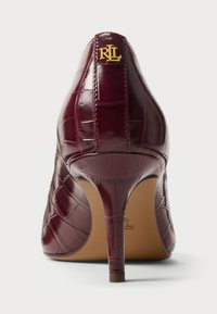 Burgundska visokotrenažna čevlja iz glatke usnjene kože s krokodilskim uzorkom, z značilnimi špičastimi prstki in zlatim logotipom na peti.