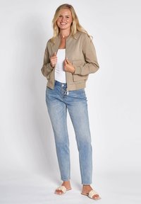 Beige Bomberjacke mit gerippten Bündchen, Reißverschluss vorne und zwei Fronttaschen, kombiniert mit hellblauen Jeans und cremefarbenen geflochtenen Sandalen.
