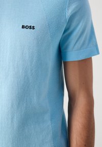 Svetlo modra kratka pletena majica s teksturiranimi navpičnimi stranskimi paneli in črnim logotipom "BOSS" na prsih, oblečena na telesu osebe.