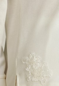 Witte geribde stof met een geborduurd bloemenontwerp en pailletten in de linker bovenhoek. Gladde textuur, slim fit.