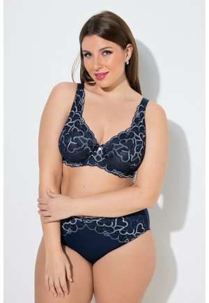 Kvinde med langt brunt hår iført navy blå bh og matchende højtaljede undertøj med blonder og blomsterprint, stående mod en hvid baggrund.