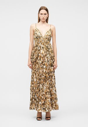 Γυναίκα που στέκεται φορώντας μακρύ, πλισέ φόρεμα με λεοπάρ σχέδιο και λεπτές ράντες, καθώς και μαύρα ψηλοτάκουνα πέδιλα με ανοιχτά δάχτυλα, πάνω σε λευκό ομοιόμορφο φόντο.