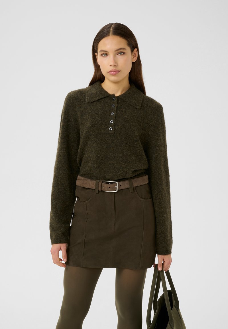 Olivgrüner Pullover mit Kragen und Knopfleiste, kombiniert mit einem braunen Wildleder-Minirock mit Taschen und einem Gürtel. Hält eine passende Tasche.