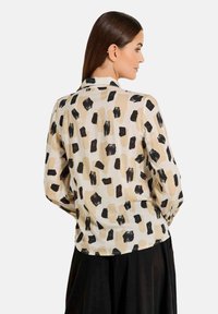 Langarmhemd aus cremefarbenem Stoff mit abstraktem schwarzen und beige Pinselstrichmuster. Kragen und leicht geschwungener Saum. Glatte Textur.