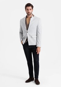 Blazer grigio chiaro con un design del colletto unico, abbinato a un gilet marrone e jeans scuri. Tessuto liscio, stile minimalista e nessun motivo visibile.